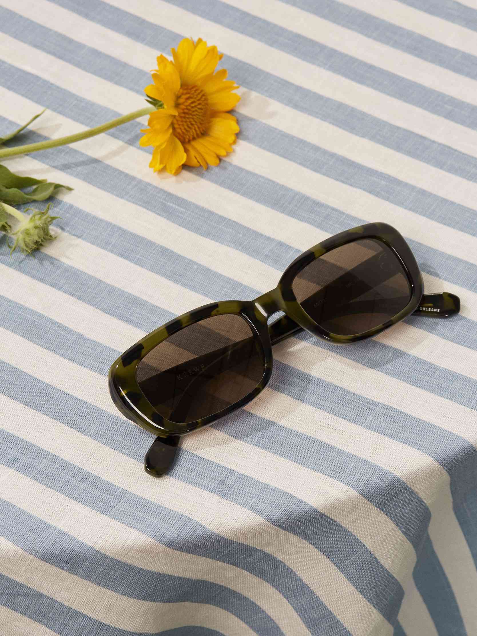 一副绿玳瑁太阳镜放在有一朵花的桌子上，还有蓝白条纹的桌布。