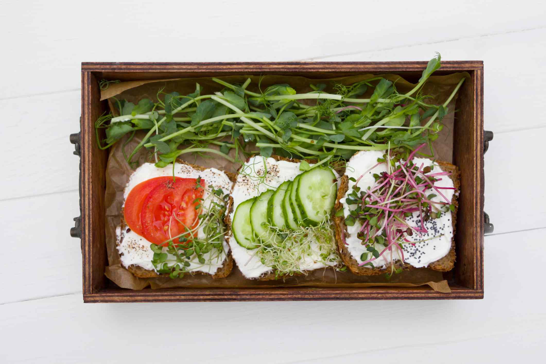 木制的盒子里放着三片吐司，奶油干酪，西红柿，黄瓜和旁边有豌豆苗的绿叶菜